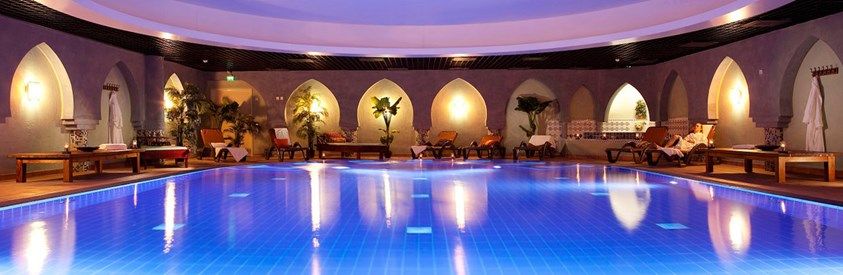 Blå upplyst inomhuspool med arabiska bågar och loungemöbler.